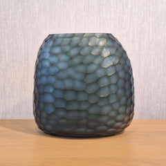Fjord Vase 24cm in deep teal-blue frosted glass, its entire surface covered in dense small hammered cells that scatter light like sunlight breaking across cold, mineral-rich water.