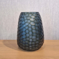 Fjord Vase 24cm in deep teal-blue frosted glass, its entire surface covered in dense small hammered cells that scatter light like sunlight breaking across cold, mineral-rich water.