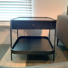 A two-tier side table with a drawer, featuring a black powder coated metal frame and a gray oak veneer tabletop with solid oak edging.