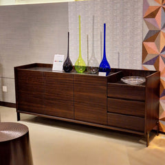 A dark brown Mora Sideboard with three doors on the left side and three drawers on the right side, made of smoked eucalyptus veneer on mdf with a glass top and stainless steel base.