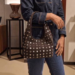 A person wearing a dark denim jacket and jeans carries a Studded Bag Suede Dark Brown with stud detailing and single top handle.