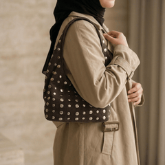 A woman wearing a beige trench coat and black hijab carries a Studded Bag Suede Dark Brown, shown from a side profile against a neutral indoor background.