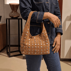 A person wearing a dark denim jacket and jeans carries a Studded Bag Suede Light Brown with stud detailing and single top handle.
