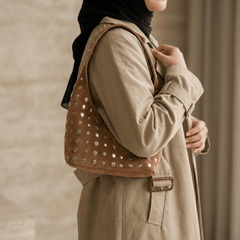 A woman wearing a beige trench coat and black hijab carries a Studded Bag Suede Light Brown, shown from a side profile against a neutral indoor background.