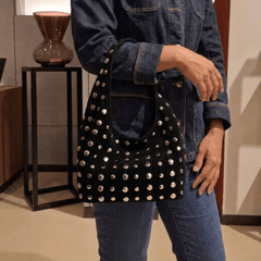 A person wearing a dark denim jacket and jeans carries a Studded Bag Suede Matte Black with stud detailing and single top handle.