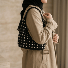 A woman wearing a beige trench coat and black hijab carries a Studded Bag Suede Matte Black, shown from a side profile against a neutral indoor background.