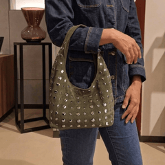 A person wearing a dark denim jacket and jeans carries a Studded Bag Suede Olive Green with stud detailing and single top handle.