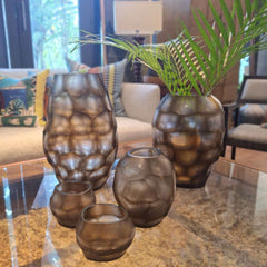 Sunhive Tealight Holder 8cm, Sunhive Vase 16cm, Sunhive Vase 27cm, Sunhive Vase 33cm in warm smoky taupe frosted glass, its surface covered in large rounded organic cells that ripple upward like honeycomb lit from within.