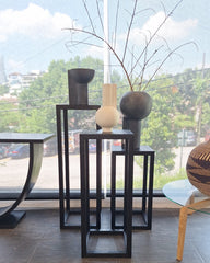 A set of modern solid ash charcoal finish Tipis Pedestal Small Ash, Tipis Pedestal Medium Ash & Tipis Pedestal Large Ash with vases arranged in front of a large window with a view of trees and sky. 