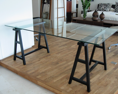 A writing table with a clear tempered glass top and charcoal finish solid ash legs, with an angled design.