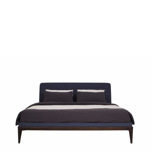 Upholstered king bed with a dark blue denim fabric headboard and footboard, placed on a light coffee colored wooden frame, and fitted with a mattress.
