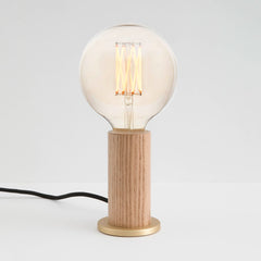 A cylindrical oak wood table lamp base with a brass in-line dimmer, featuring a smooth finish and a tactile dimmer switch, by Tala
