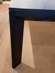 A modern rectangular dining table made of solid ash with a charcoal finish, featuring a mitre joint at the corners and stained legs.
